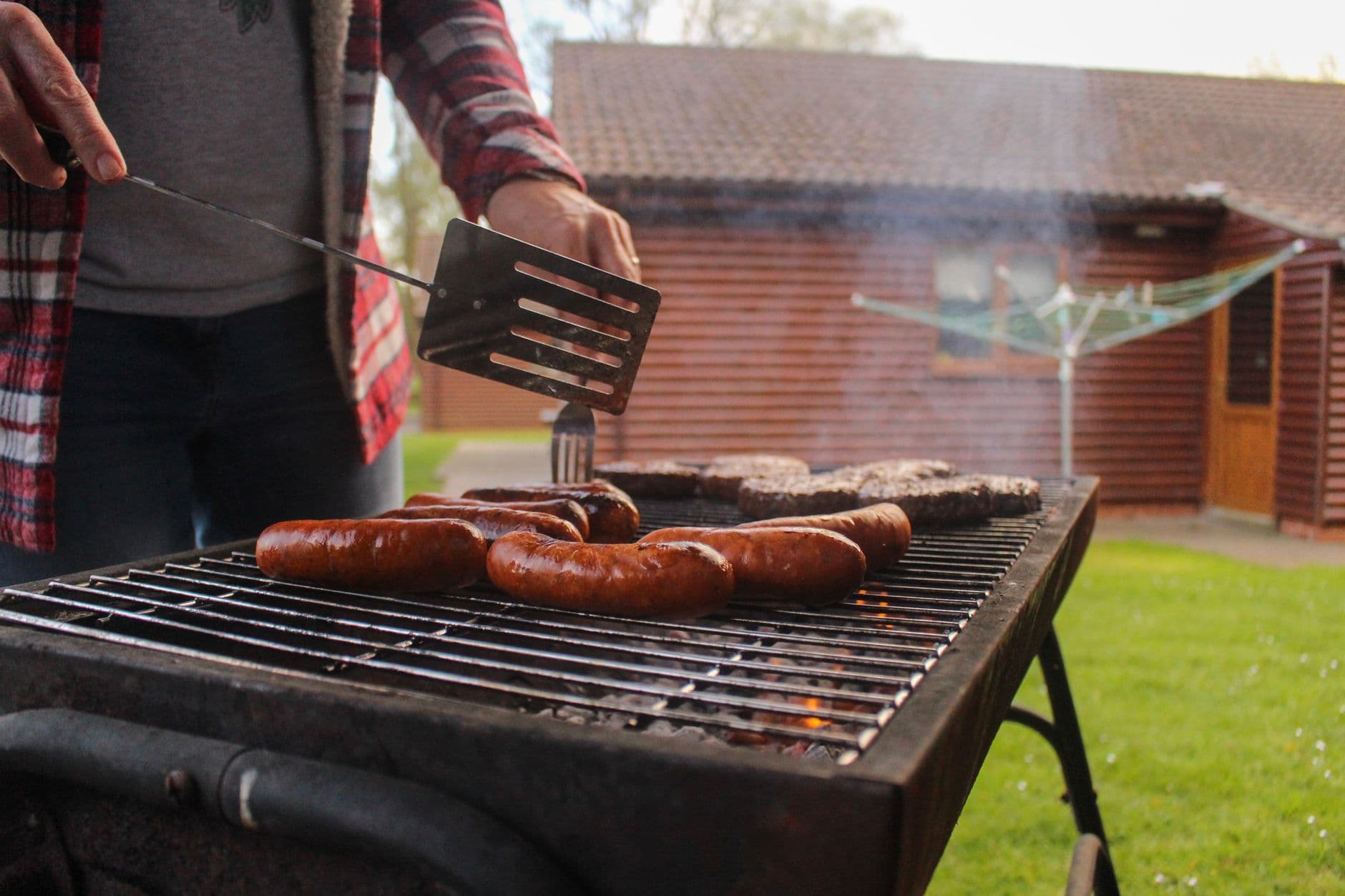 Grilling sausages and patties in a backyard with a rustic wooden house in the background.